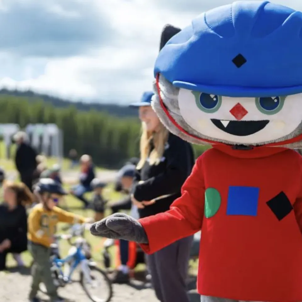 Ein Cartoon-Maskottchen mit blauem Helm und rotem Hemd steht im Vordergrund; Kinder und Erwachsene sind in der Parkumgebung dahinter zu sehen.